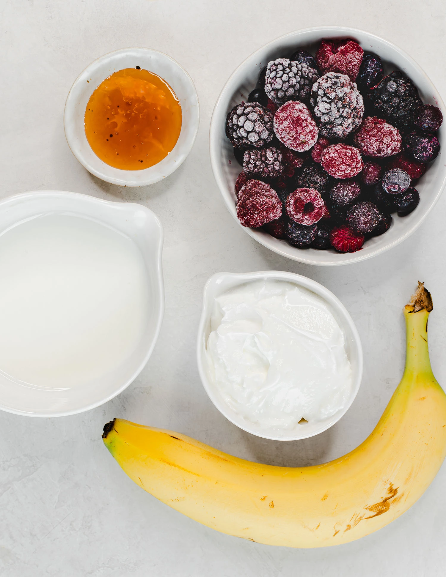 Yogurt, whole milk, honey, and frozen mixed berries including raspberries, blueberries, and blackberries separated into bowls on a countertop next to a banana.