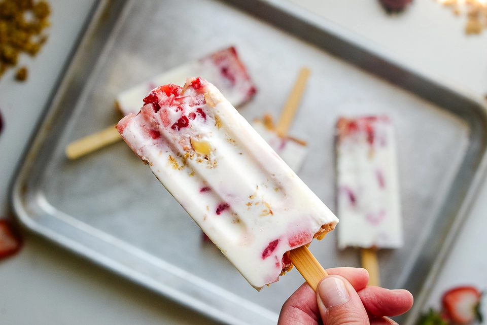 Hand holding a single frozen strawberry Greek yogurt popsicle, showing visible layers of red strawberries, thick white Greek yogurt, and toasted oats.