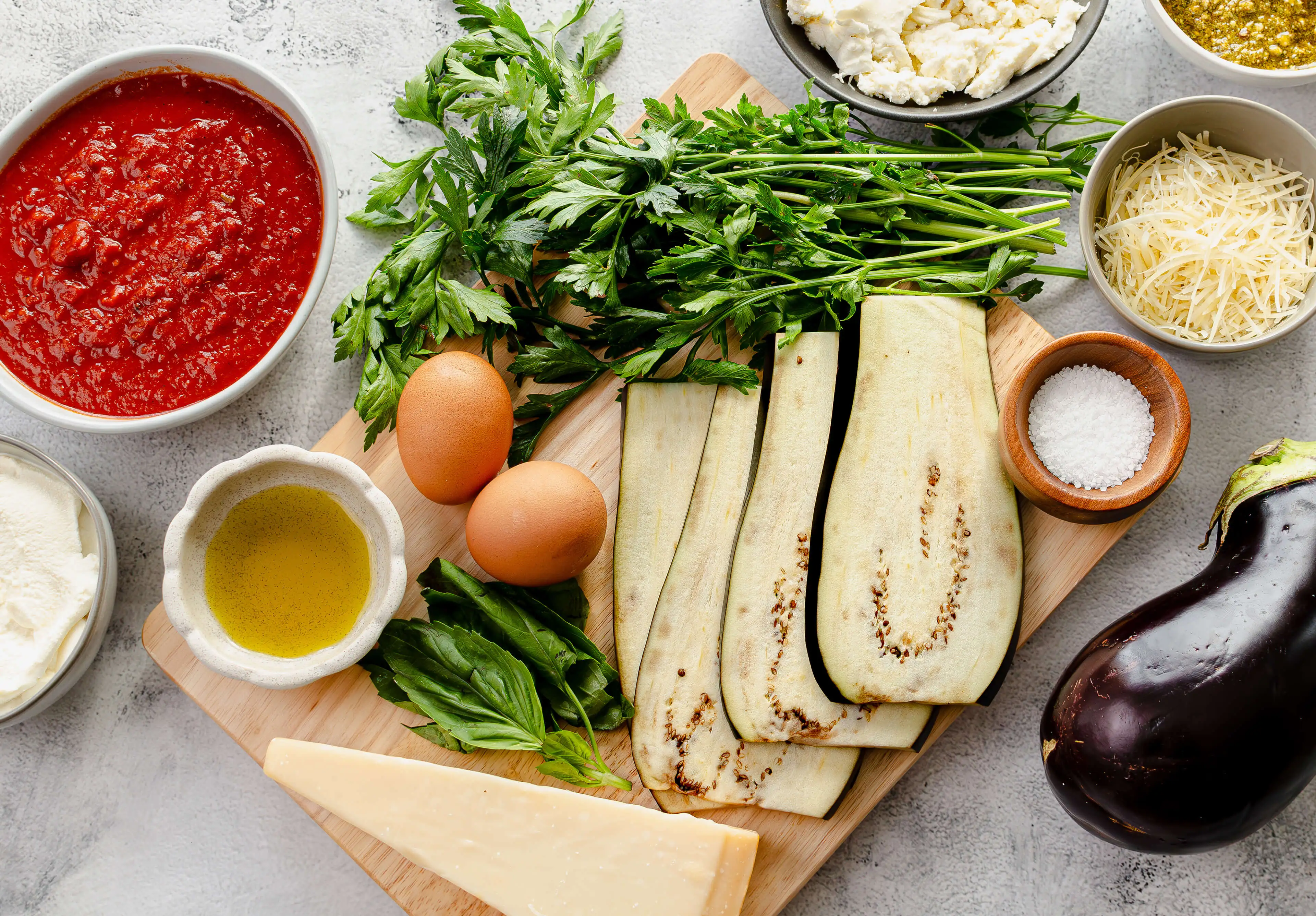 Ingredients for an eggplant rollatini such as eggplant, marinara sauce, parmesan, mozzarella, ricotta, basil and parsley on a cutting board.