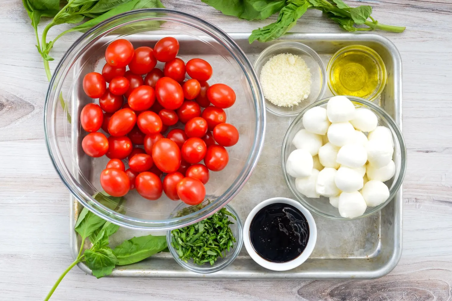 Ingredients for a caprese salad wreath appetizer on a tray, such as: tomatoes, basil, mozzarella cheese, olive oil, Parmesan cheese, and a balsamic glaze