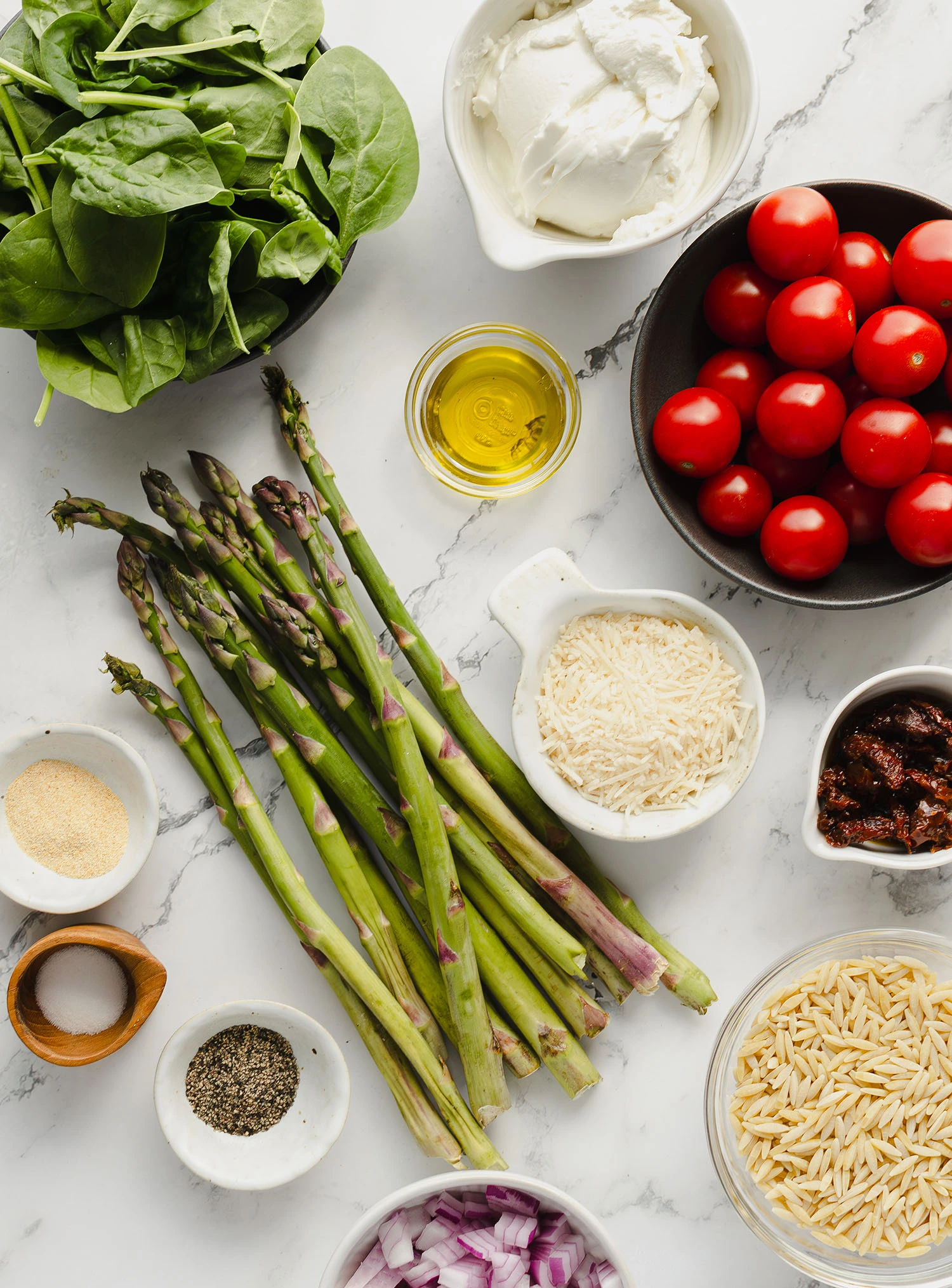 Breakdown of ingredients from tomato, asparagus, spinach, orzo, and cheese to spices like salt, pepper, and garlic powder measured in bowls on a marble countertop