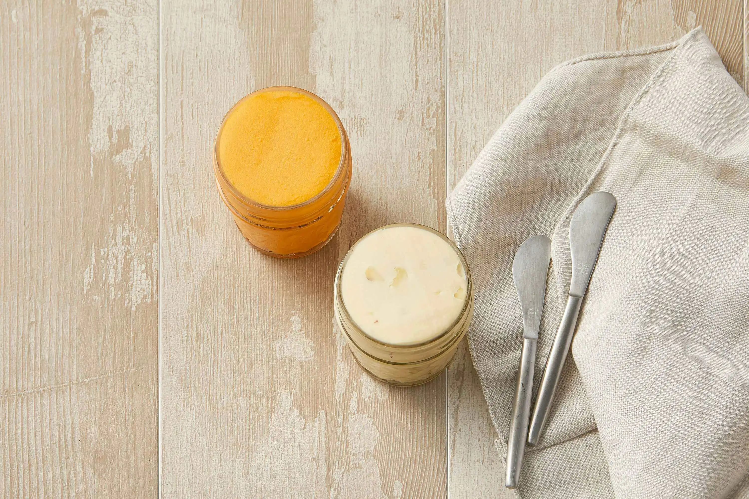 Two glass jars filled with a luscious creamy cheese spread, accompanied by two silver knives and a folded linen napkin.
