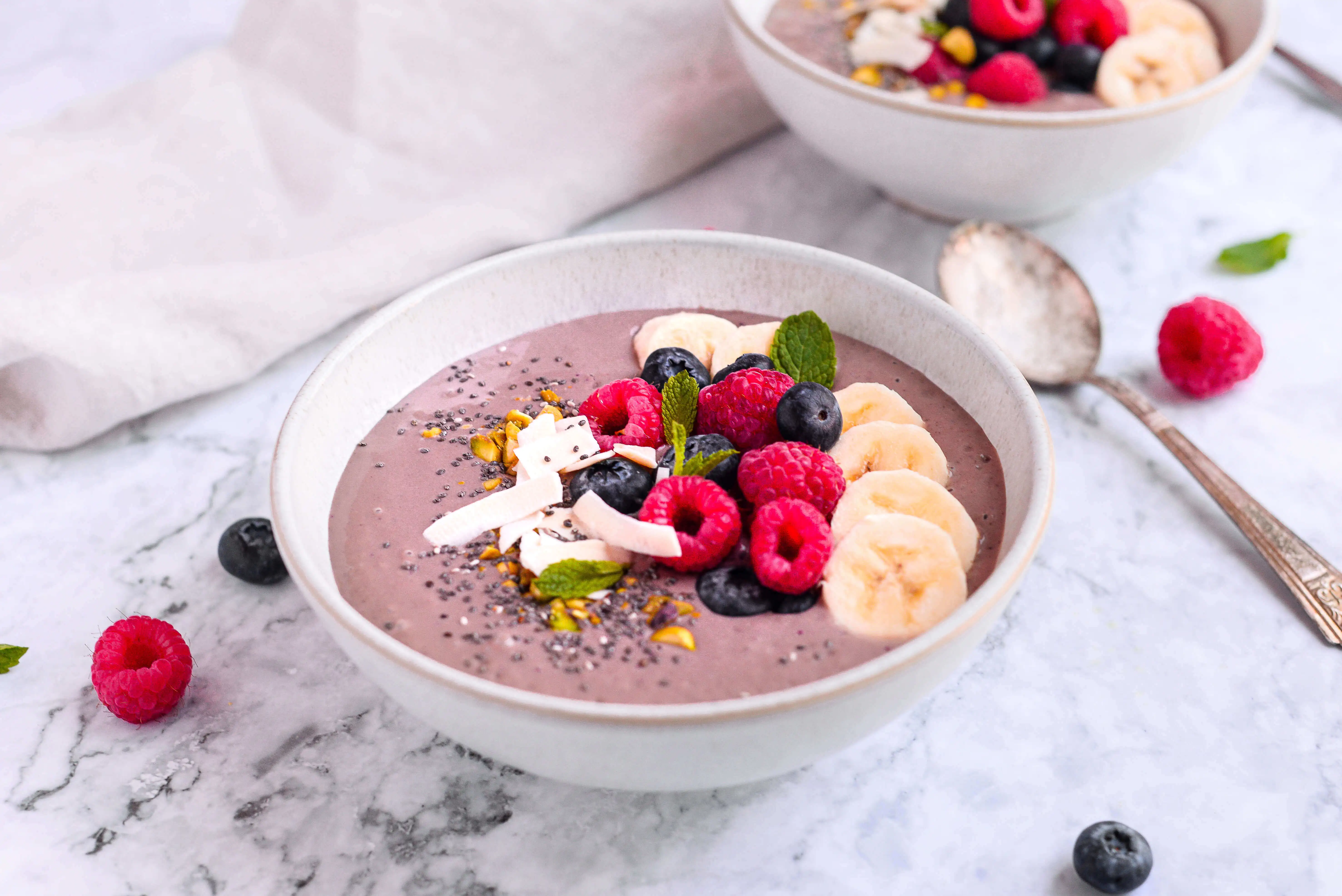 Acai bowl topped with berries, chia seeds, coconut chips, pistachios and mint sitting on a table with a spoon.