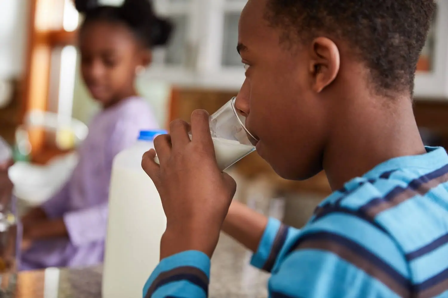 child drinks glass of milk with a pitcher and another kid in the background