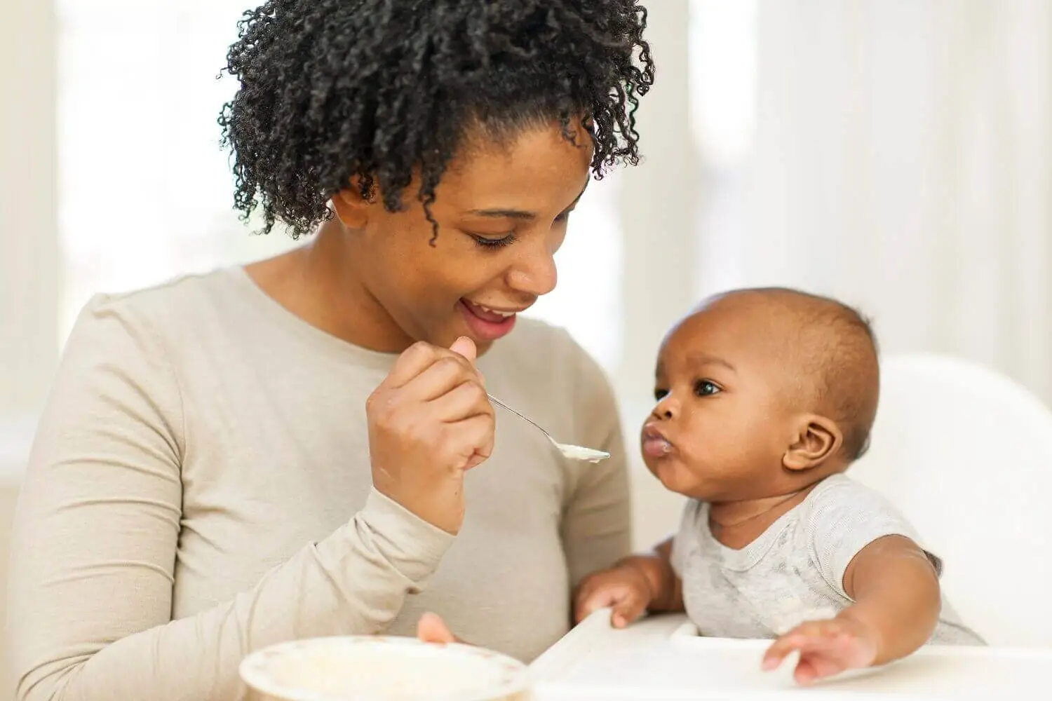 A mother, holding a spoon and plate, feeds her baby seated in a high chair.