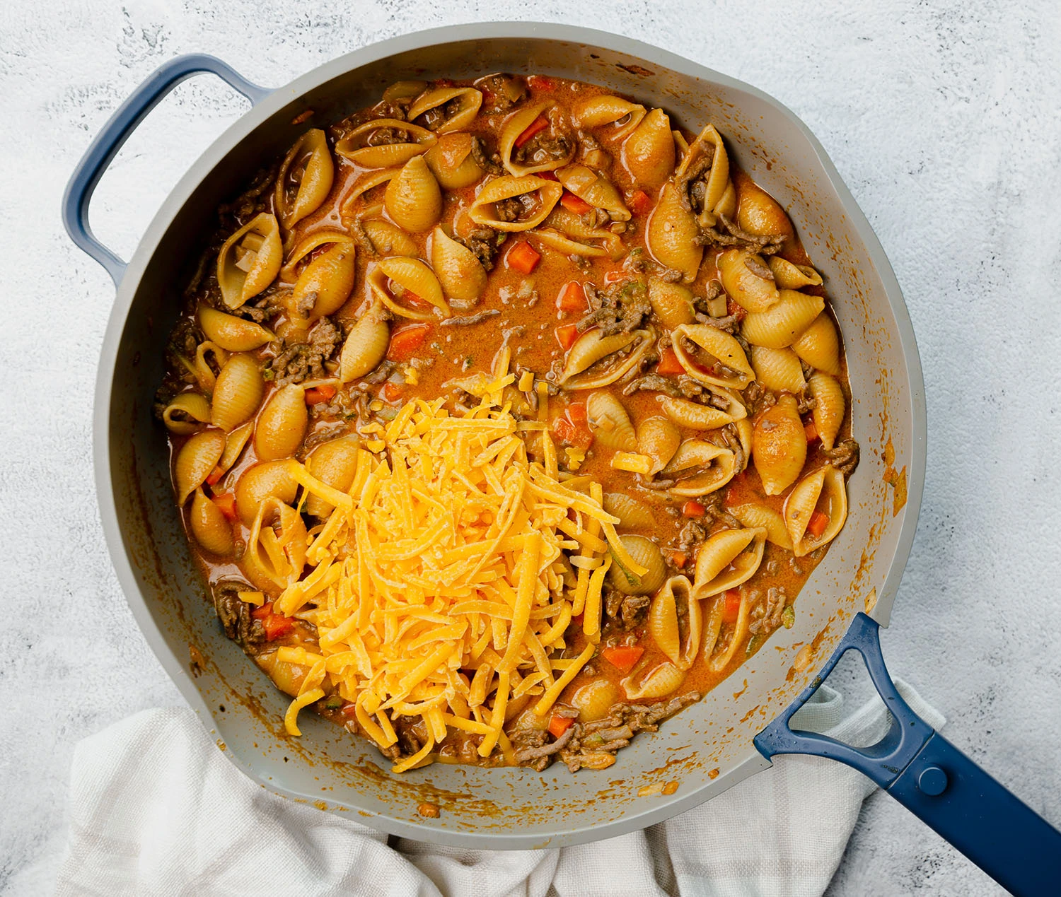 A pan filled with cooked pasta, ground beef, and vegetables assortment including carrots, onions, mushrooms, and zucchini with a pile of cheddar cheese on the top.