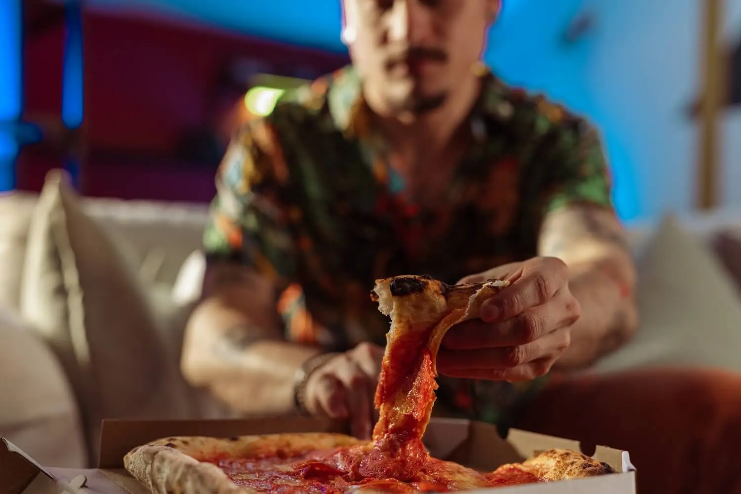 A man sitting in an armchair, reaching for a slice of pizza from the box.