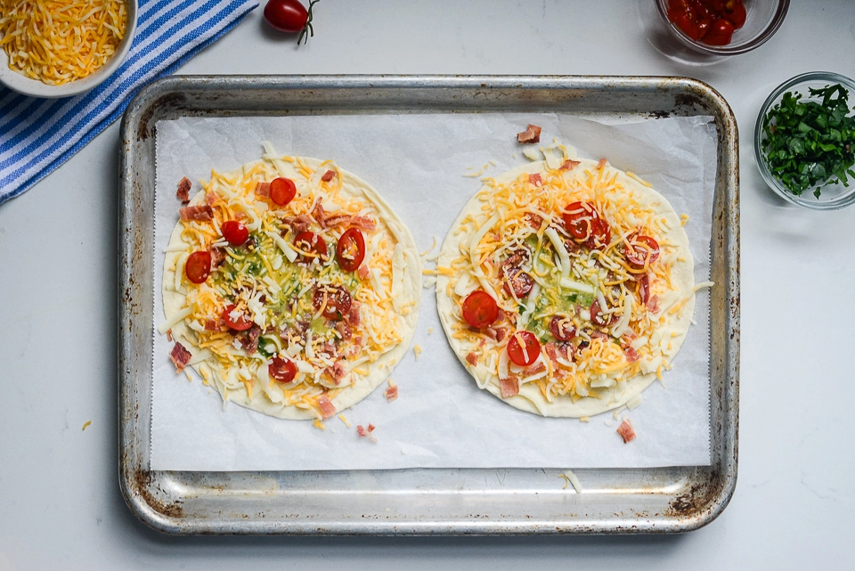 Unbaked breakfast pizzas topped with cheese, bacon, tomatoes, and egg on a parchment-lined baking sheet.