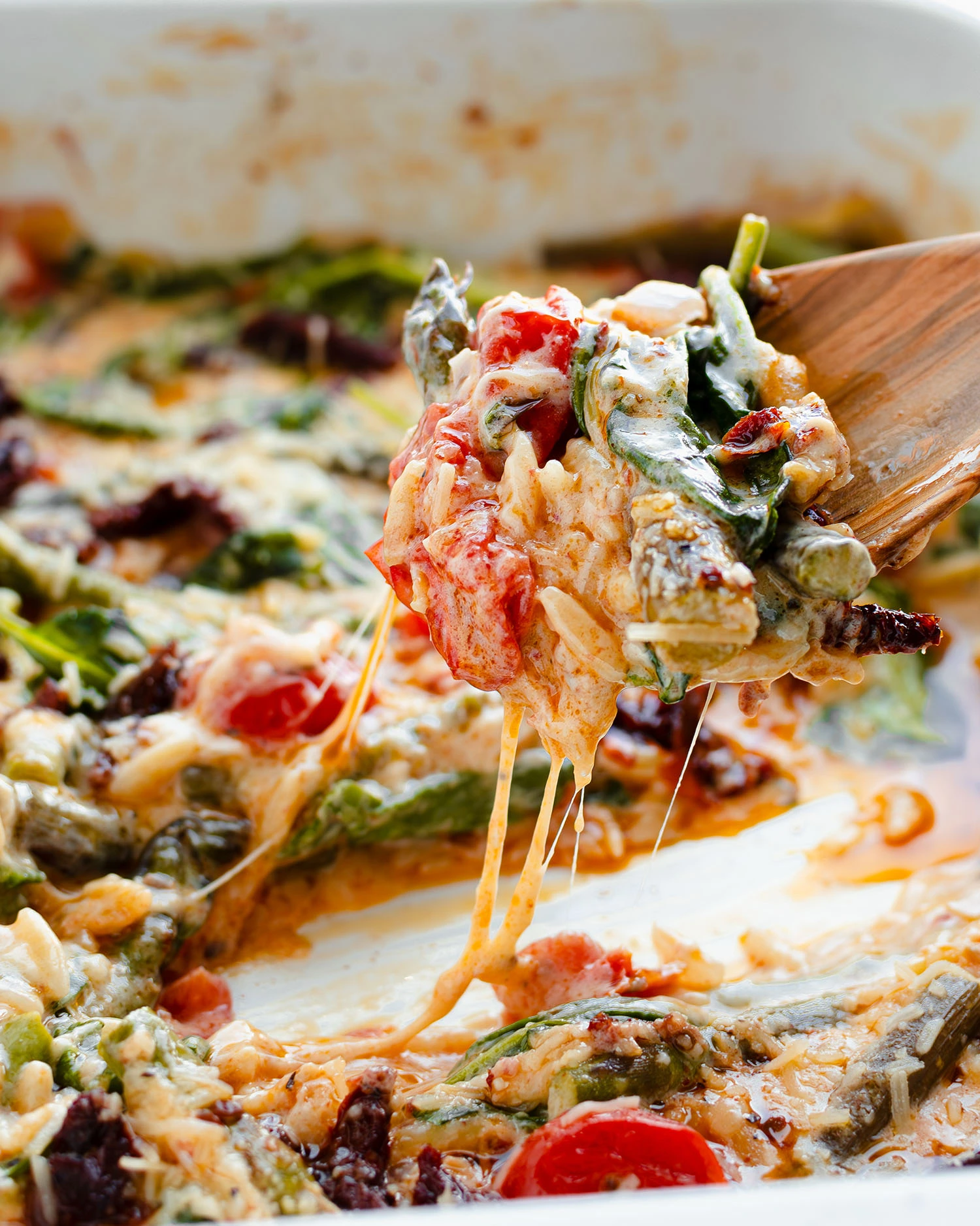 A bite of the roasted orzo bake topped with gooey cheese on a serving spoon displayed over a baking dish.