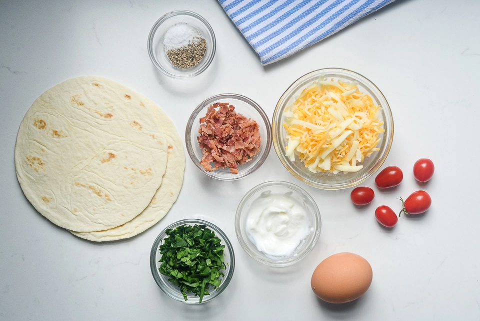 Overhead view of ingredients for crispy bacon, egg & cheese breakfast pizza, including tortillas, bacon, cheese, spinach, yogurt, egg, tomatoes, and seasonings.