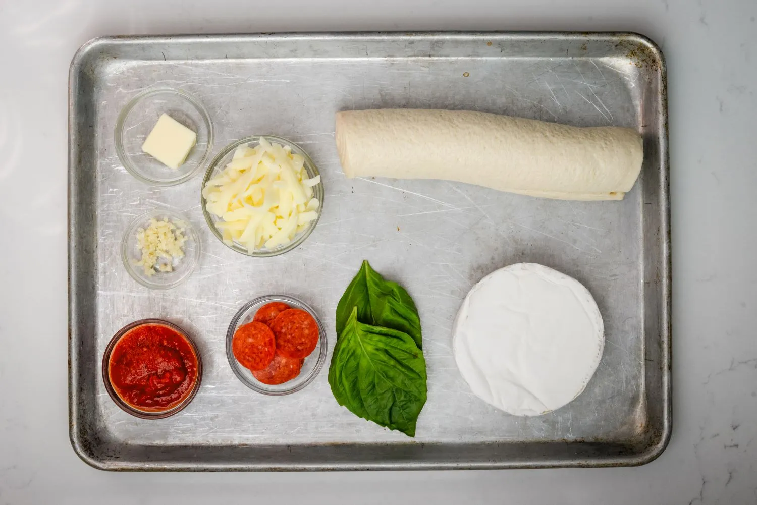 Ingredients for Brie stuffed pizza including Brie cheese wheel, pizza sauce, garlic, pepperoni, and basil