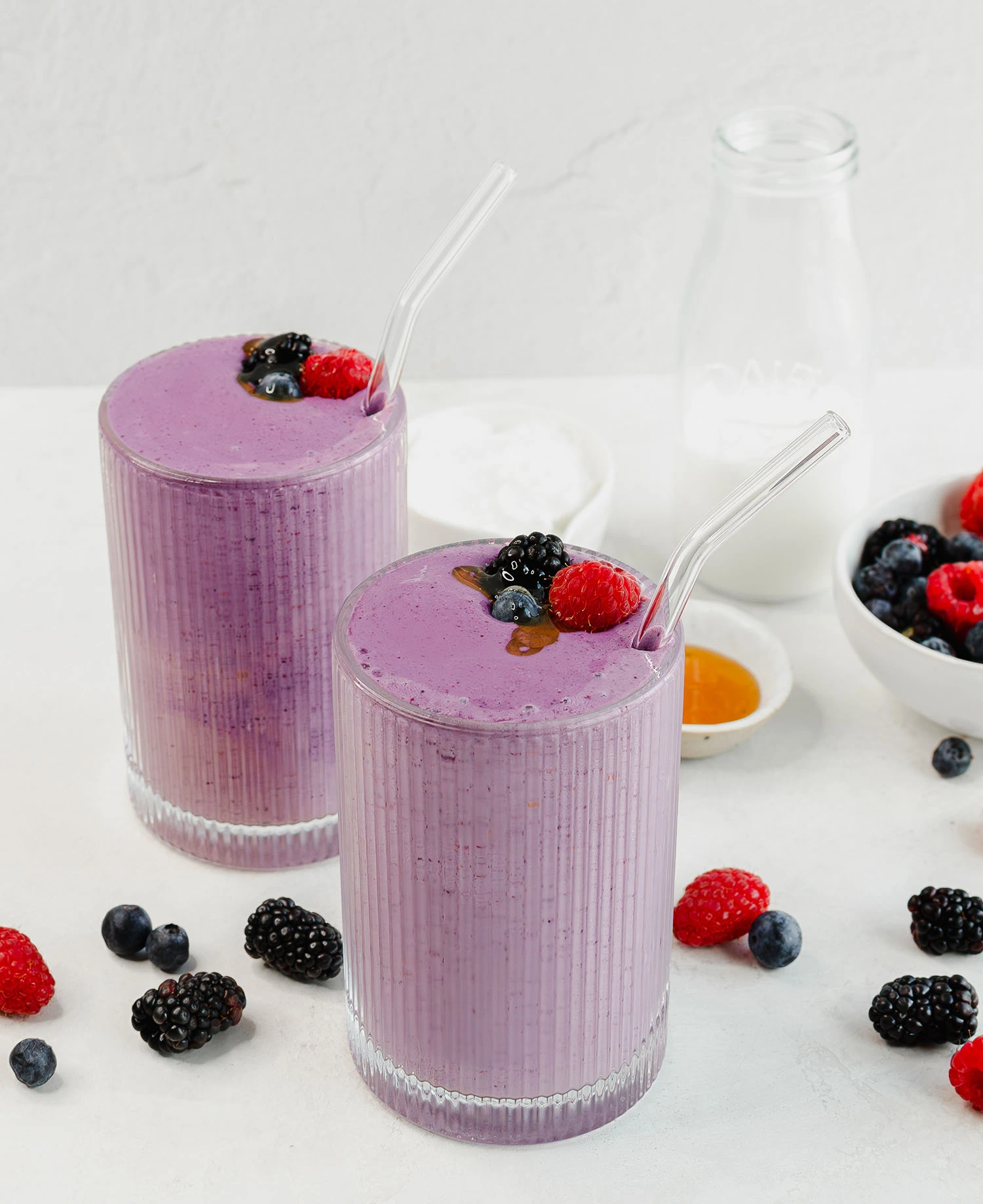Two glasses filled to the brim with a protein berry smoothie, topped with raspberries, blueberries, and blackberries with extra supply of ingredients in the background including mixed berries and milk