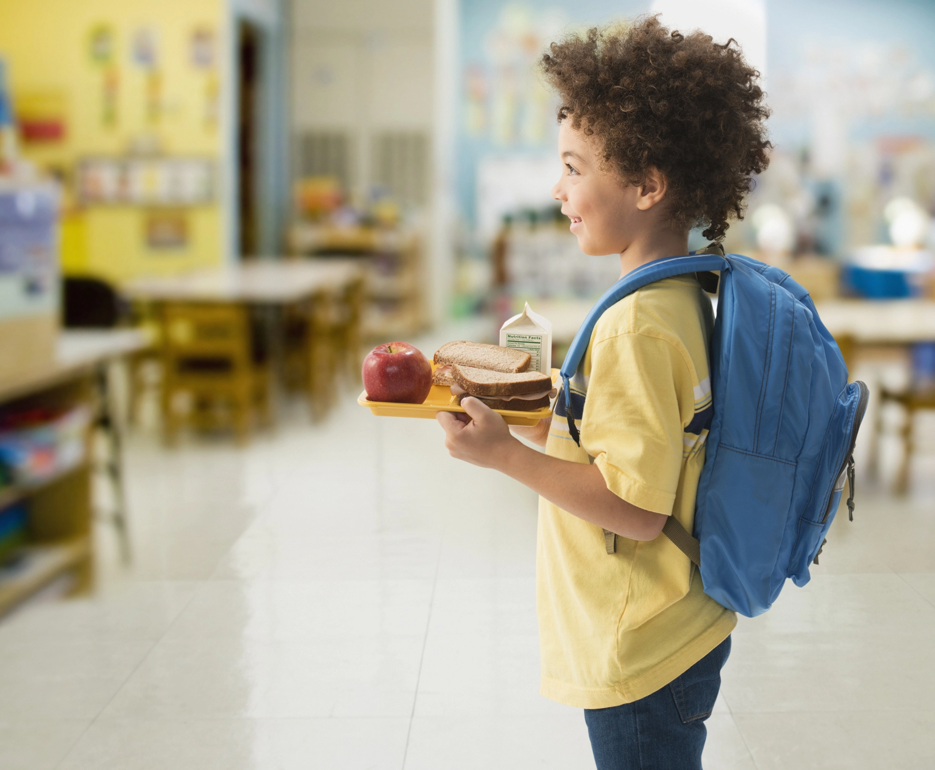 A child at school holding a lunch tray with a sandwich, apple, and carton of milk.