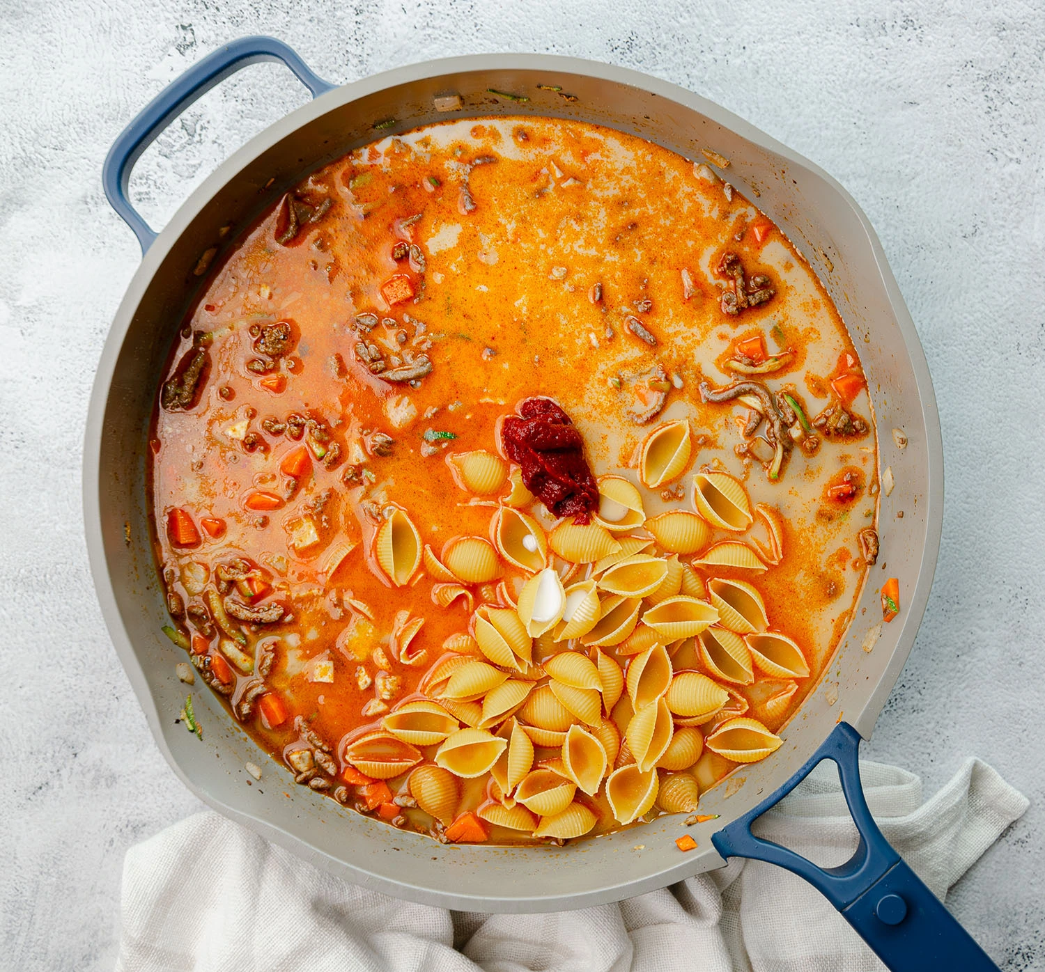 A pan of uncooked pasta shells with ground beef and vegetable mixture of zucchini, onions, carrots, and mushrooms, covered in broth and milk combined with tomato paste.