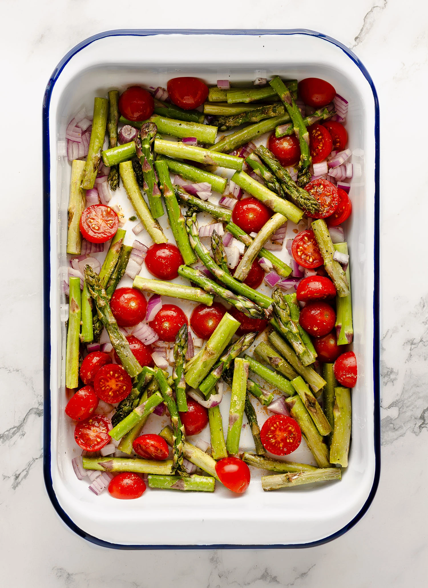 Seasoned asparagus, onions, and tomatoes with olive oil in an enamel baking pan on a countertop.