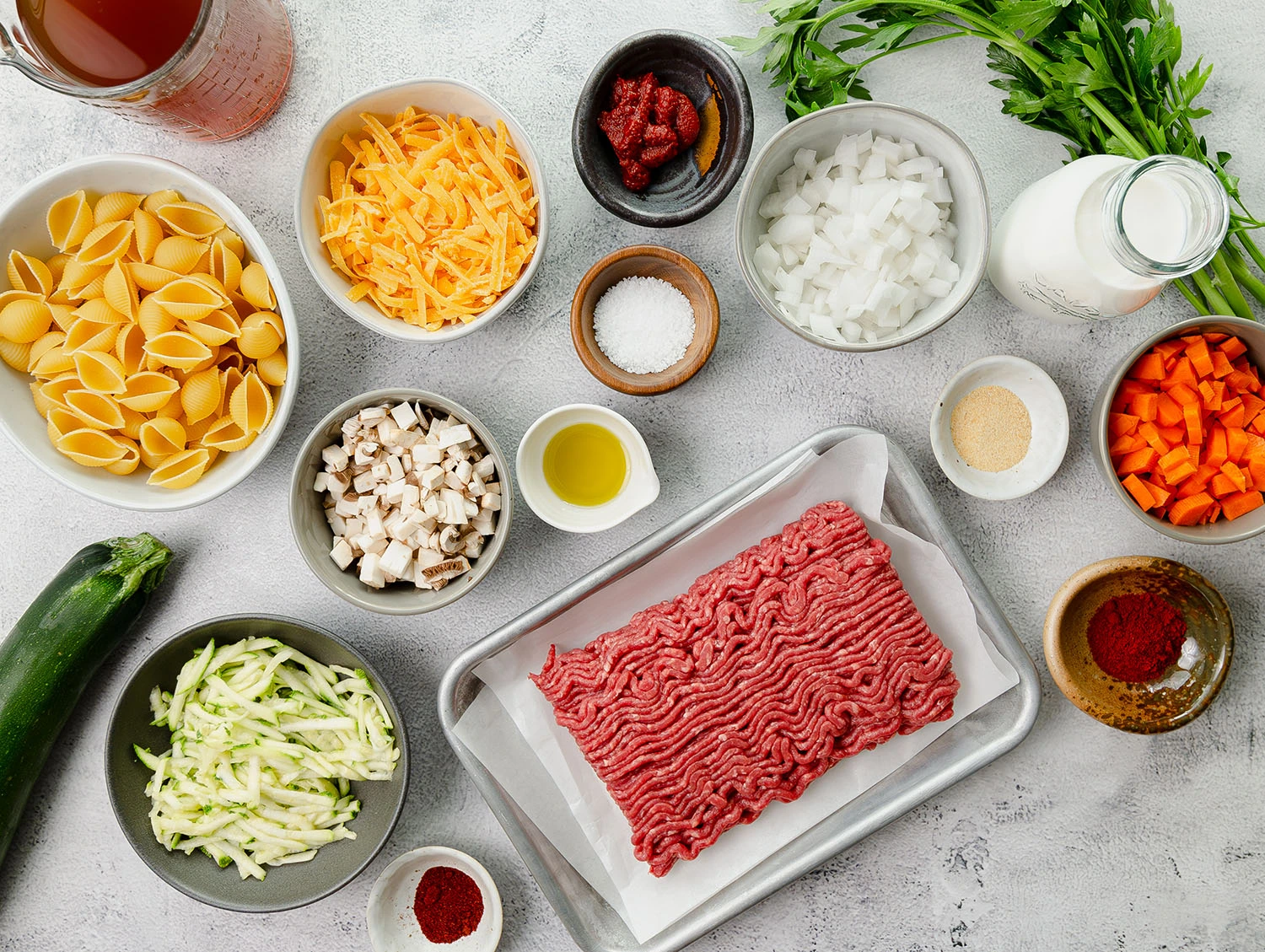 A variety of ingredients for a hamburger pasta recipe such as ground beef, pasta, cheese, vegetables, seasonings, and milk are laid out on a counter-top.